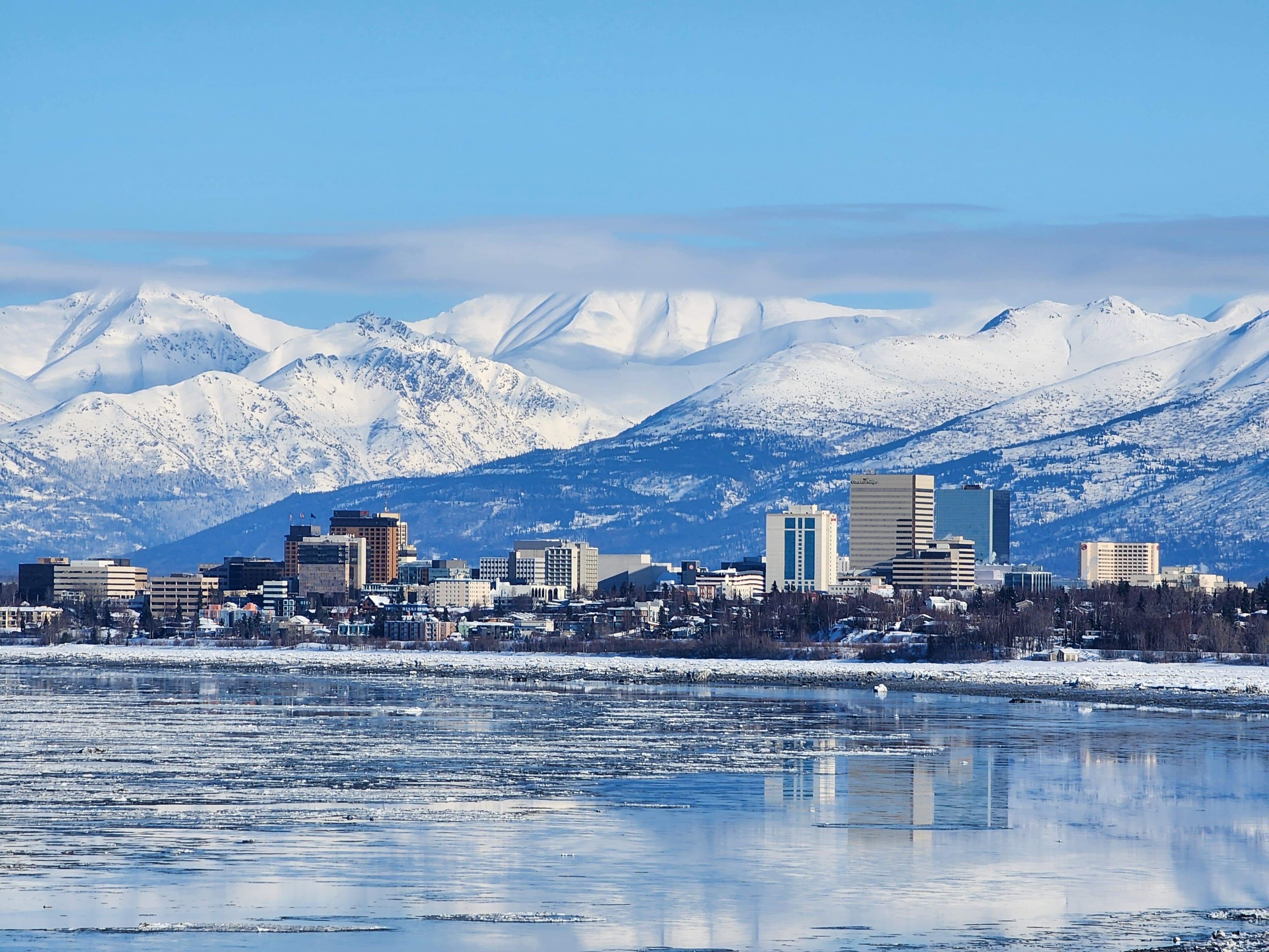 A stunning view of Alaskan city surrounded by snow-covered mountains, with a serene lake reflecting the clear sky above. The landscape is adorned with winter scenery and buildings nestled against the backdrop of majestic peaks.
