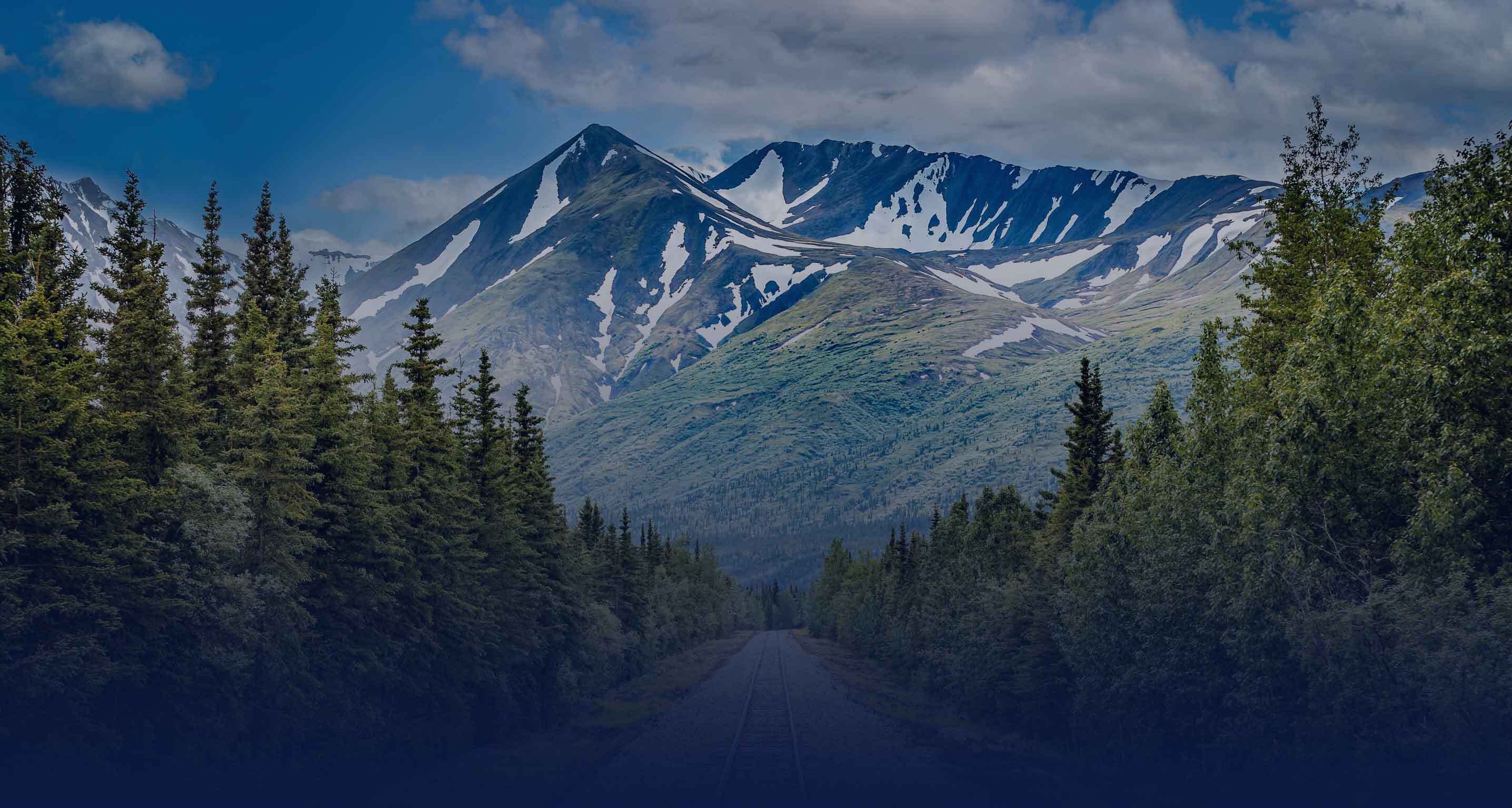 A serene lake is nestled among lush green trees and towering mountains, reflecting the sky above with scattered clouds.