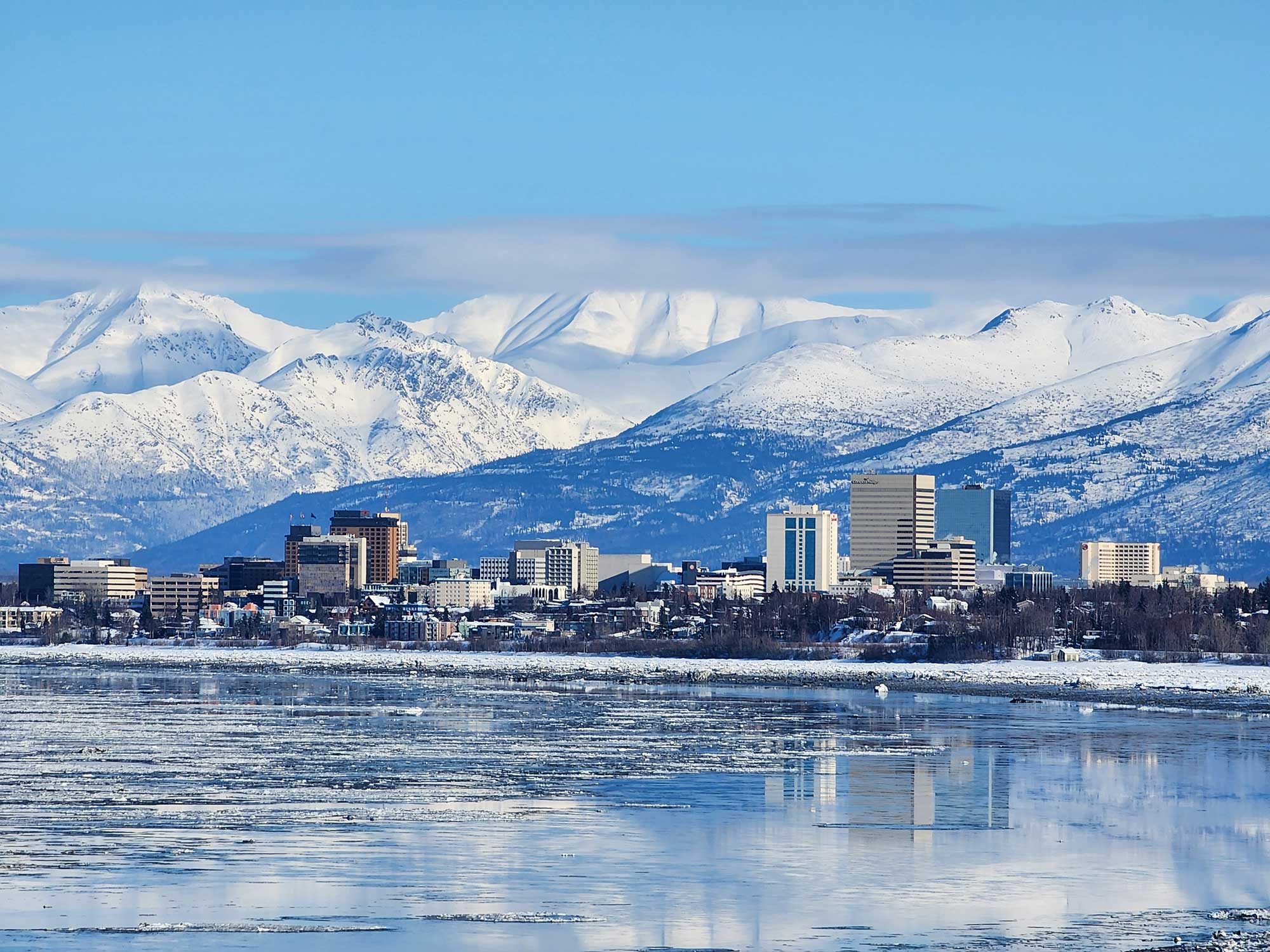 Alaskan city is situated next to a body of water, with snow-covered mountains in the background. The scene includes clear skies, reflecting the winter landscape.