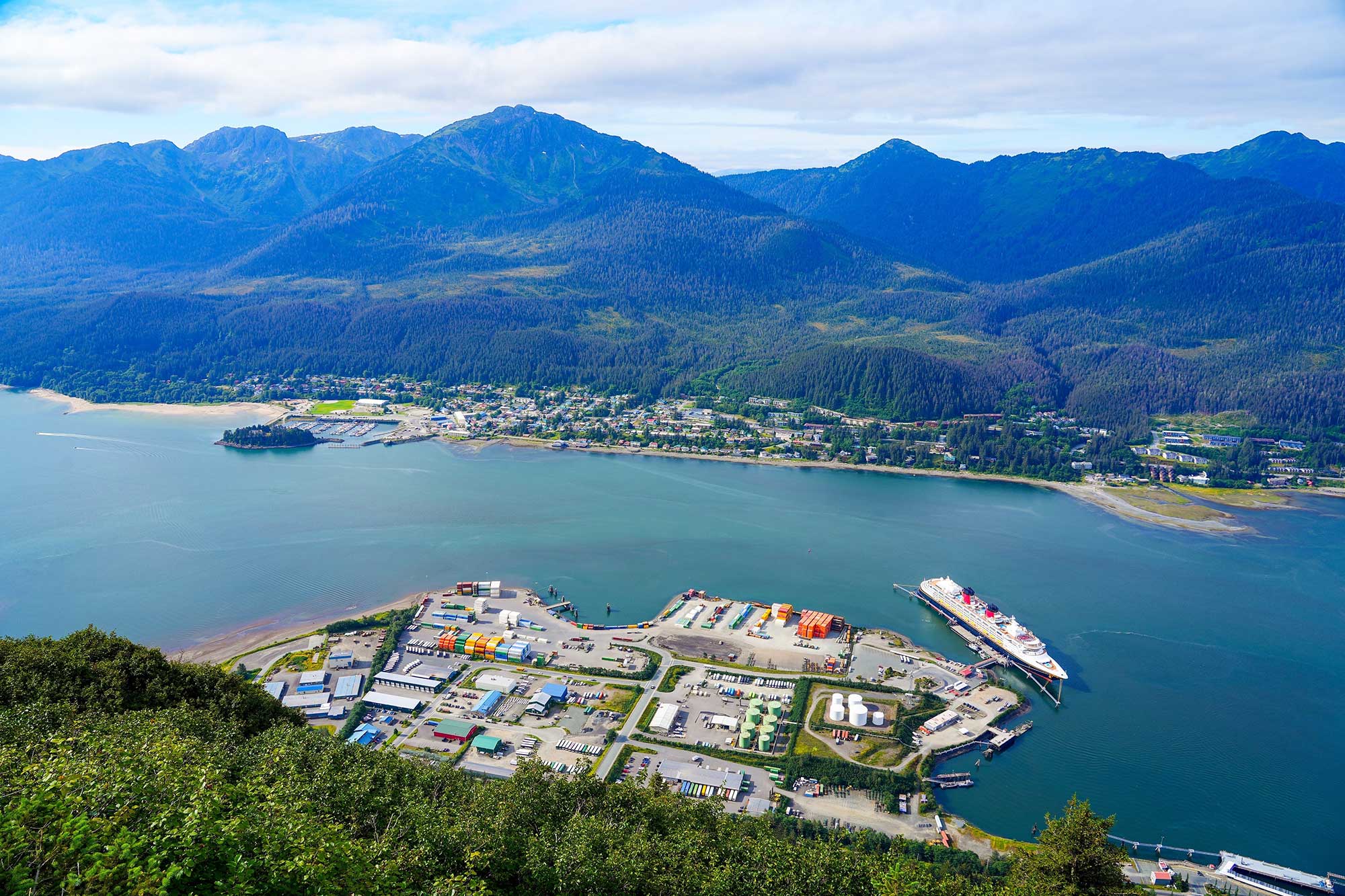 Aerial view of a Alaskan city adjacent to a body of water, featuring lush green trees and mountainous terrain in the background. The sky is partly cloudy, highlighting the natural landscape that includes an inlet or lake district.