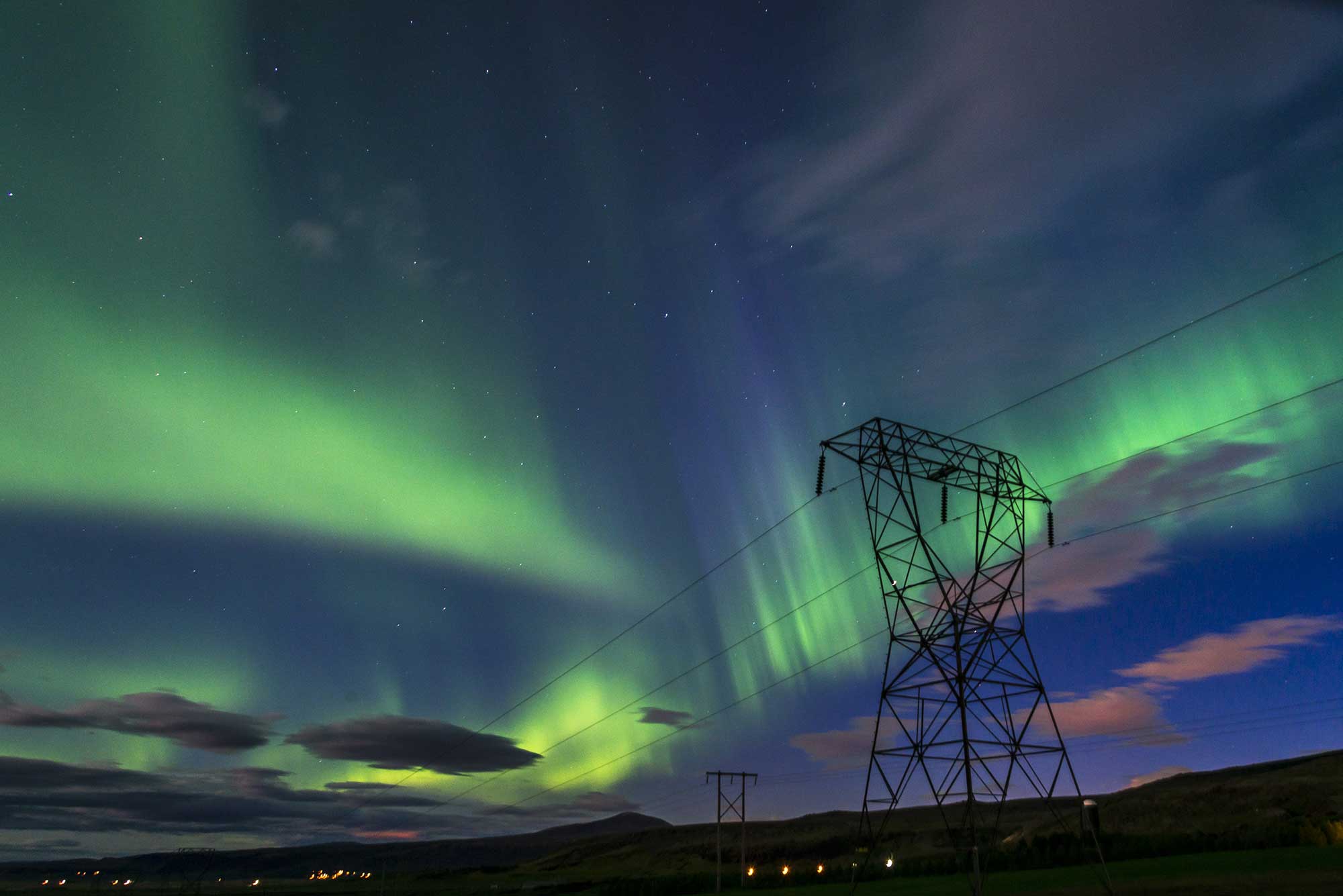 A tall tower illuminated by colorful lights against a night sky filled with clouds and an aurora, surrounded by green grass.